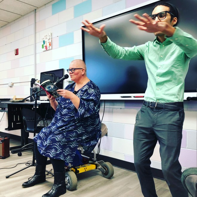 In the foreground, a man wearing green provides ASL interpretation for Petra Kuppers' reading of Ice Bar. Petra, a white woman wearing blue glasses and a blue dress covered in lighter blue fronds, speaks into a microphone.
