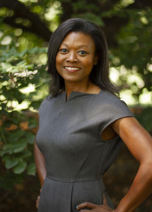 A Black woman stand in front of out-of focus greenery. She has long black hair and is wearing an inviting smile and shot-sleeved black dress.