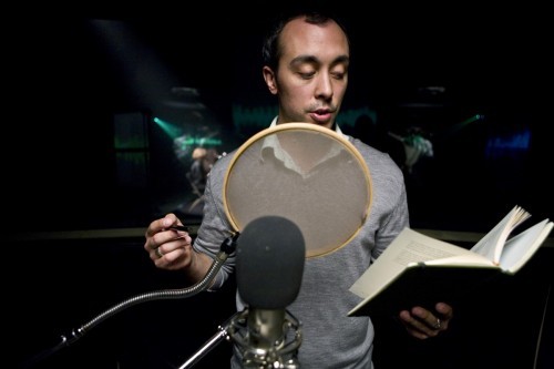 A white man with short black hair is reading aloud behind a pop filter and microphone. He is looking at a book in his left hand.