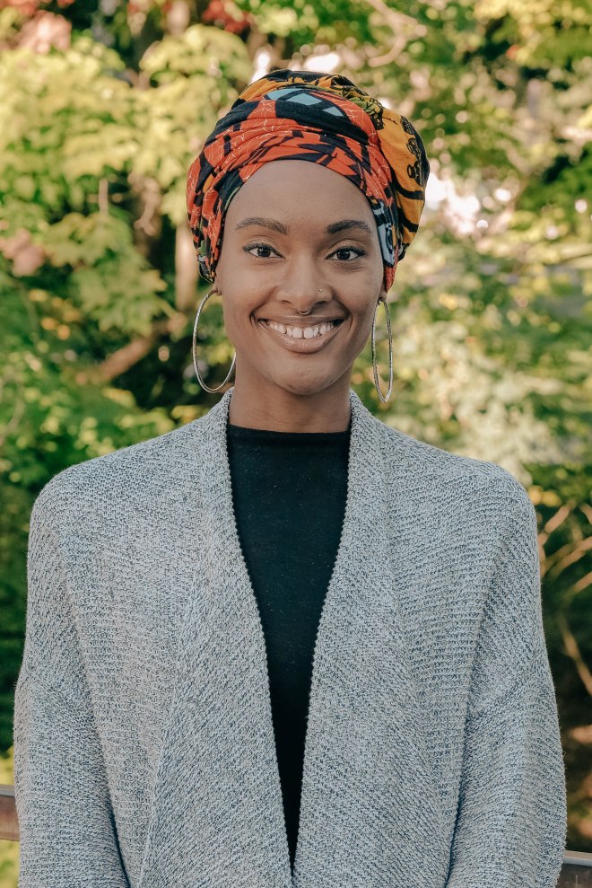 A woman with Black skin stands for a professional photograph. She is wearing a fine gray cardigan over black clothing and an orange, teal, and black head wrap. In the background is out of focus greenery.