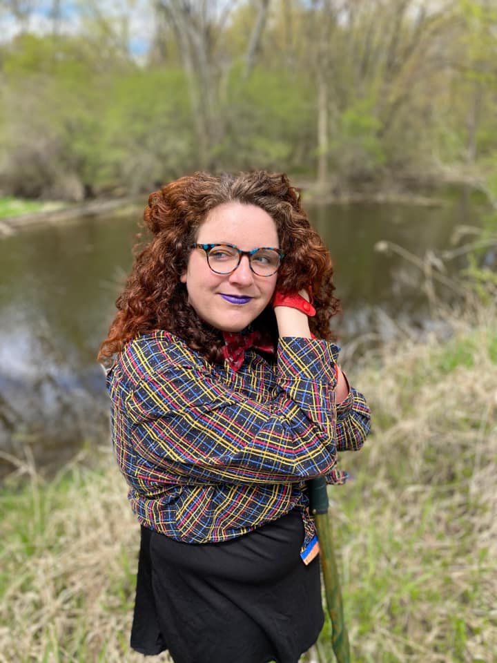 A person with a lot of red, curly hair and white skin is wearing purple lipstick. They are wearing a button up shirt criss-crossed with red, blue, and yellow on a on a starry black background. They lean on a green walking stick.