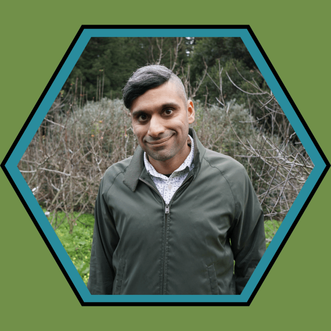 Dr. Neel Ahuja standing in front of green bushes and trees. He has brown skin and short brown hair. He wears a green jacket over a white button-down shirt with black patterning.