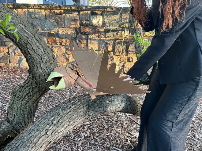 A bird made of recycled cardboard rests atop a tree. In the background are woodchips, a brick wall, and an early spring sky. The cardboard bird is puppeteered by a person wearing all black clothing who has red hair. No skin or identifying features of the puppeteer are visible.